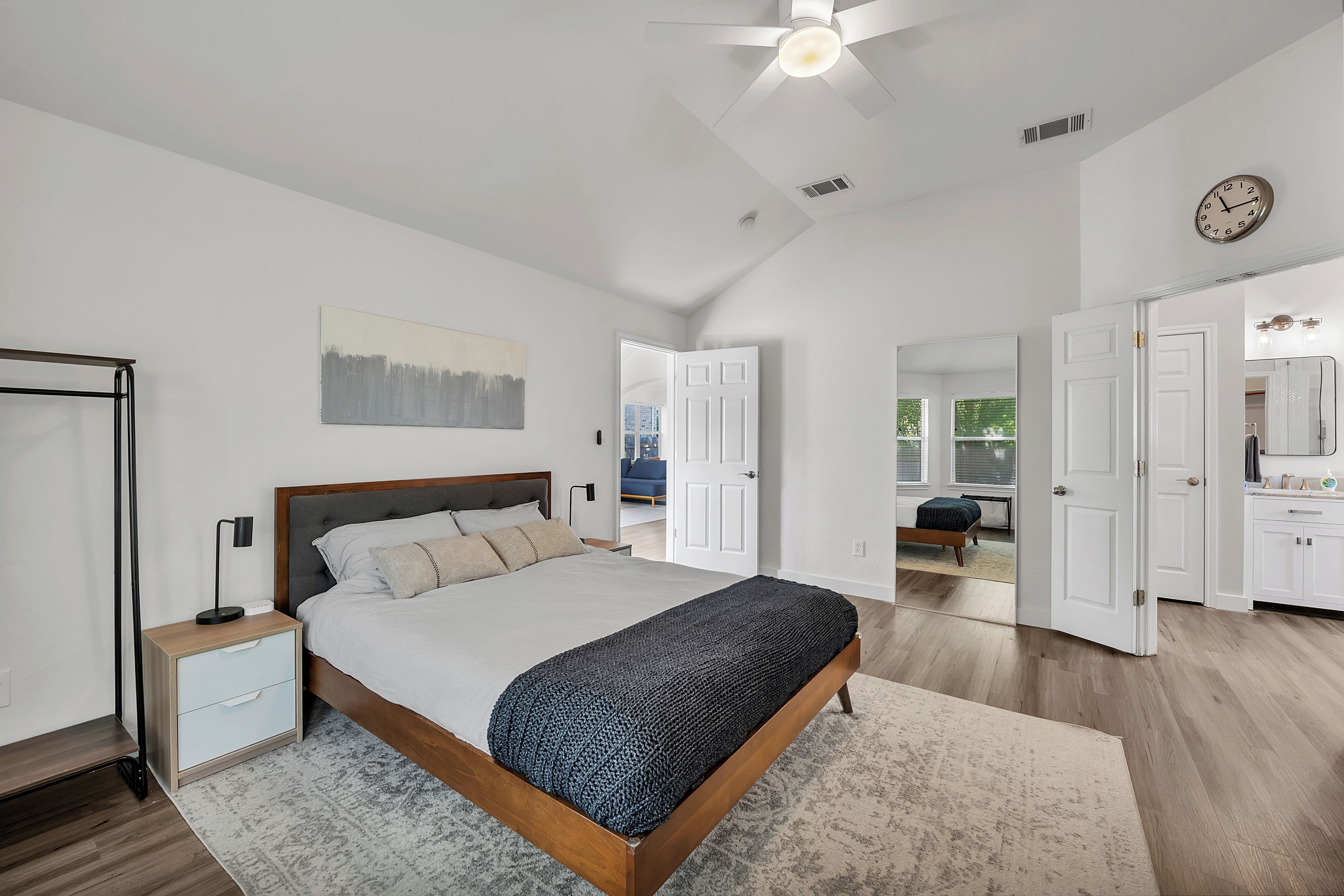 A bedroom with a bed, bedside tables, a ceiling fan, a clock, and an open door leading to a bathroom.