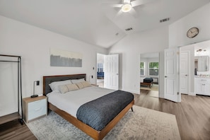 A bedroom with a bed, bedside tables, a ceiling fan, a clock, and an open door leading to a bathroom.
