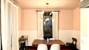 Un coin repas avec une table en bois, des chaises blanches, un lustre et une fenêtre avec des rideaux.