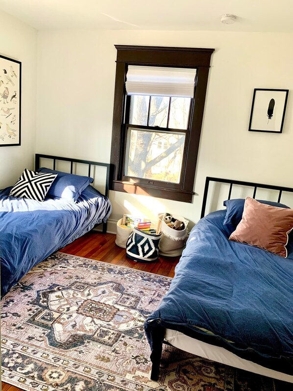 Une chambre avec deux lits, une fenêtre avec des stores, un tapis et un mur décoré de tableaux encadrés.