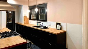 Une cuisine moderne avec un plan de travail en bois, des meubles noirs, une cuisinière à gaz, des luminaires suspendus et une fenêtre aux montants noirs.