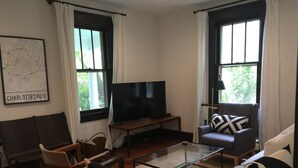 Un salon équipé d’un téléviseur à écran plat, de deux fenêtres avec des rideaux, d’une carte accrochée au mur et d’un canapé agrémenté de coussins à motifs.
