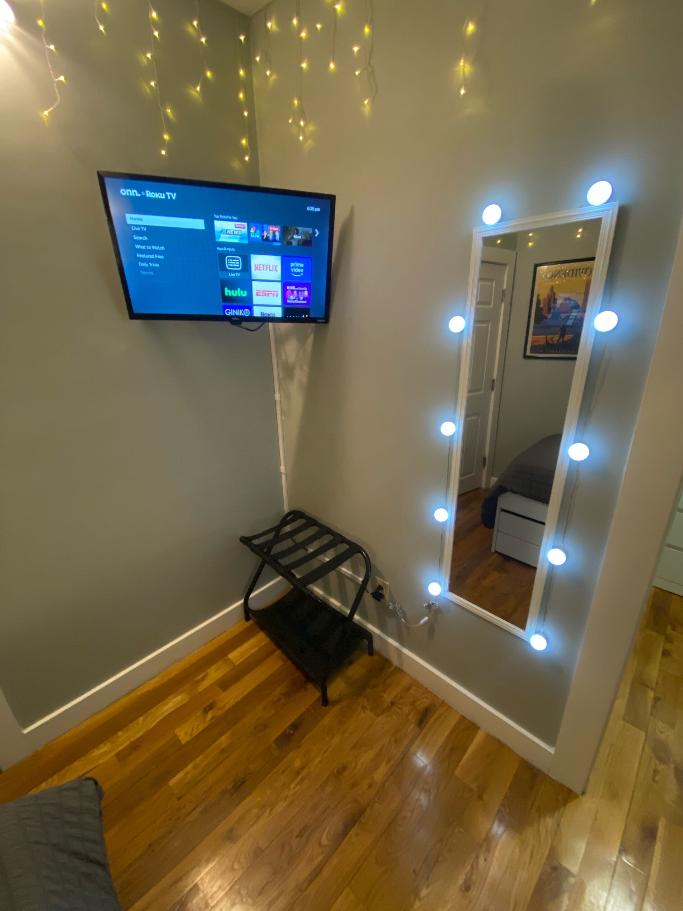 Una stanza con TV a parete che mostra un'interfaccia di servizio di streaming, uno specchio trucco con luci e un mobile contenitore nero.