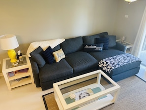 Een moderne woonkamer met een donkergrijze bank, een witte bijzettafel met een gele lamp en een glazen salontafel.