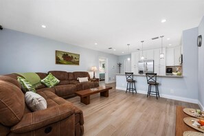Sala de estar con un sofá de cuero marrón, una mesa de centro de madera y una cocina con armarios blancos y un refrigerador de acero inoxidable.
