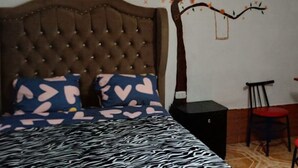 A bedroom with a tufted headboard, a zebra-patterned bedspread, heart-patterned pillows, a small black nightstand, a red chair, and a white shelving unit.