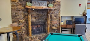 A pool table in a room with a stone fireplace, a television, and a small table.