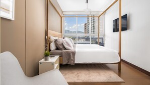Un dormitorio con cama, mesita de noche y vistas a edificios a través de un amplio ventanal.