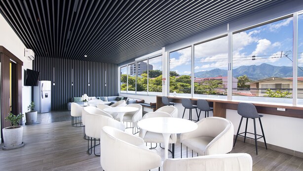 Un interior moderno con un largo pasillo que conduce a una zona de estar con mesas y sillas, amplias ventanas con vistas a edificios y montañas.