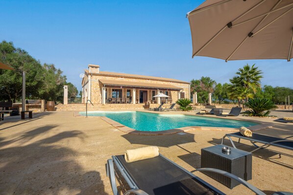 Un espace aménagé au bord de la piscine, avec des chaises longues, un grand parasol et un bâtiment à la façade en pierre.