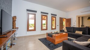 Una sala de estar moderna con una mesa de centro de madera, un sofá y un televisor sobre un mueble de madera. Hay tres ventanas con marcos de madera y una alfombra negra en el suelo.