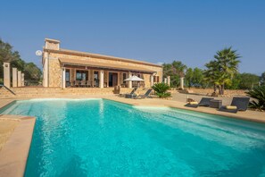 Zona de piscina amb un edifici de pedra, seients de relax i una piscina ampla i blava.