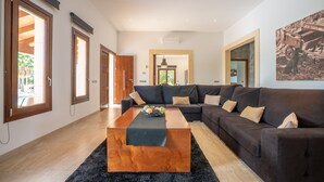 Un salón espacioso con un sofá modular grande, una mesa de centro de madera y una alfombra negra. Se observan ventanas amplias en un lateral y una puerta que da a otra habitación.