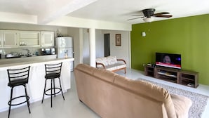 Una sala moderna con un sofá color beige, dos taburetes negros, un televisor y una zona de cocina con armarios blancos y una pared de acento verde.