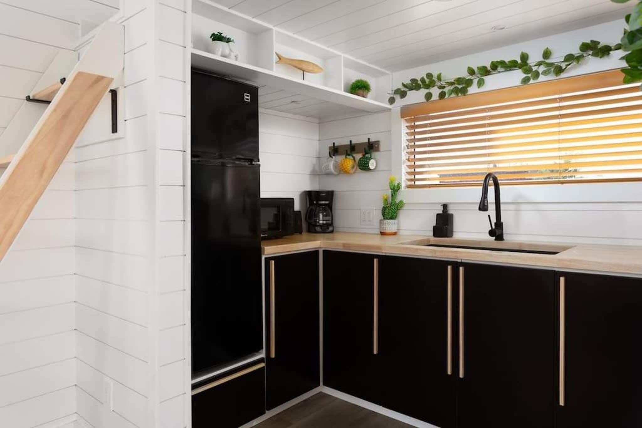 Een keuken met zwarte kasten, een zwarte koelkast, een houten aanrecht en een raam met zonwering.