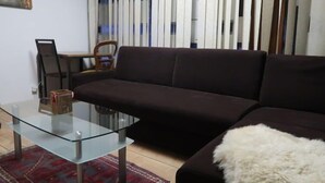 Ett vardagsrum med ett soffbord i glas, en mörkbrun soffa, en vit, fluffig pläd, en röd, mönstrad matta och en tavla på väggen.