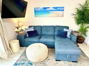 Una sala de estar con un sofá modular, un otoman redondo, un televisor de pantalla plana, un cuadro de una playa, una lámpara y una planta en maceta.