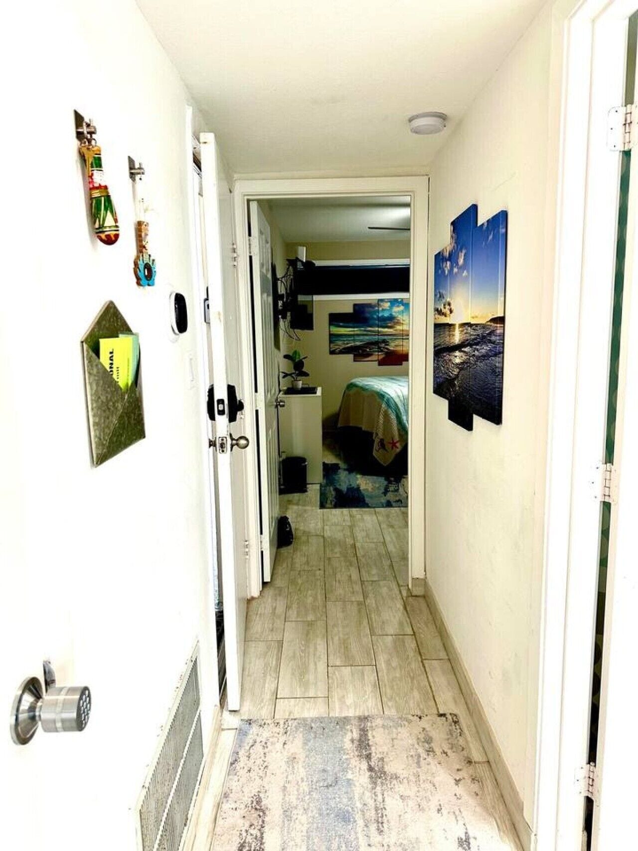 A narrow hallway with a bed in the background, a door on the left, and a wall with various decorations.