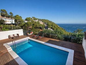 A rooftop pool with wooden deck and lounge chairs, overlooking a coastal landscape with houses and trees.