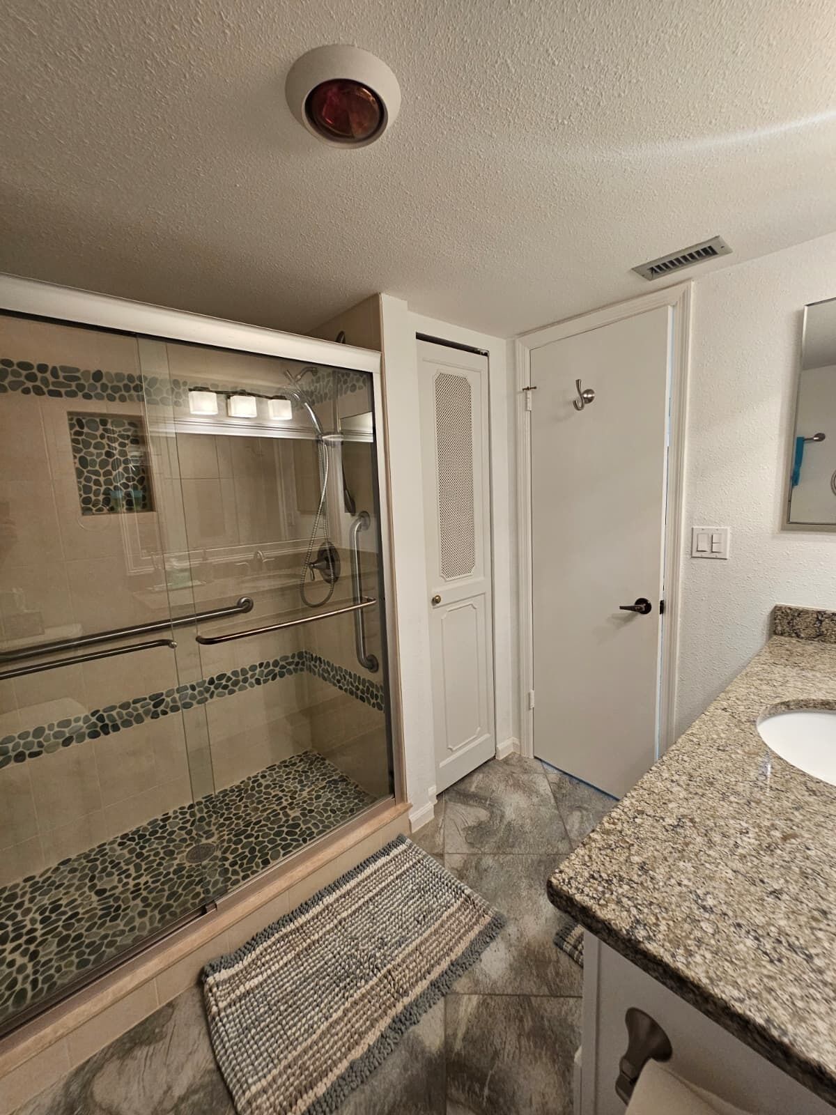 Baño con cabina de ducha de vidrio, puerta blanca, encimera de granito y suelo de baldosas.