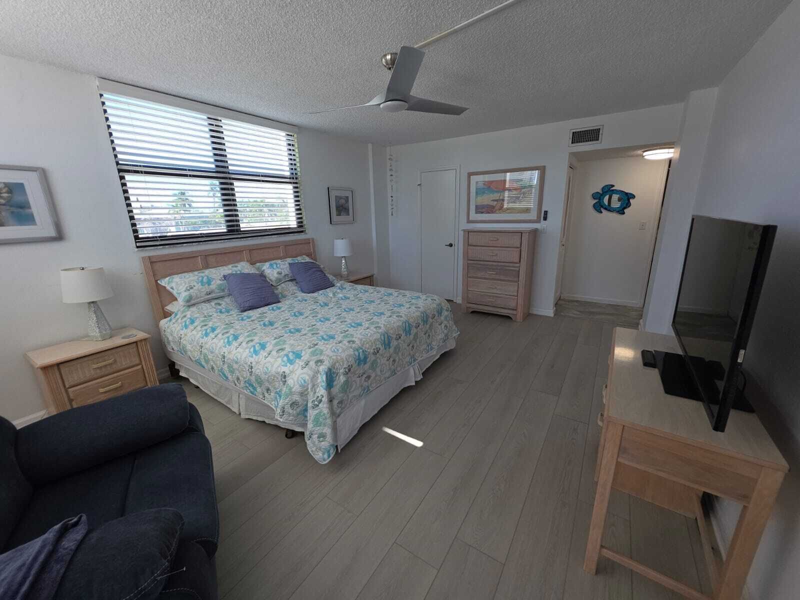 Une chambre à coucher comprenant un lit, un bureau, une chaise, un ventilateur de plafond et un téléviseur.