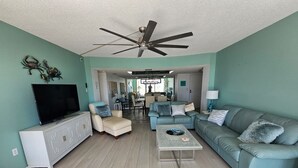 A living room with teal walls, a ceiling fan, a TV, a white cabinet, a beige armchair, a blue sofa, and a coffee table. - Large Fort Myers Beach 2 Bedroom Condo (Fort Myers Beach)
