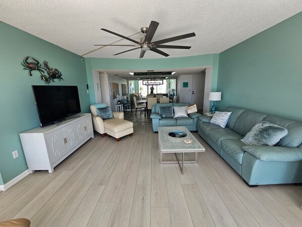A living room with teal walls, a ceiling fan, a TV, a white cabinet, a beige armchair, a blue sofa, and a coffee table.