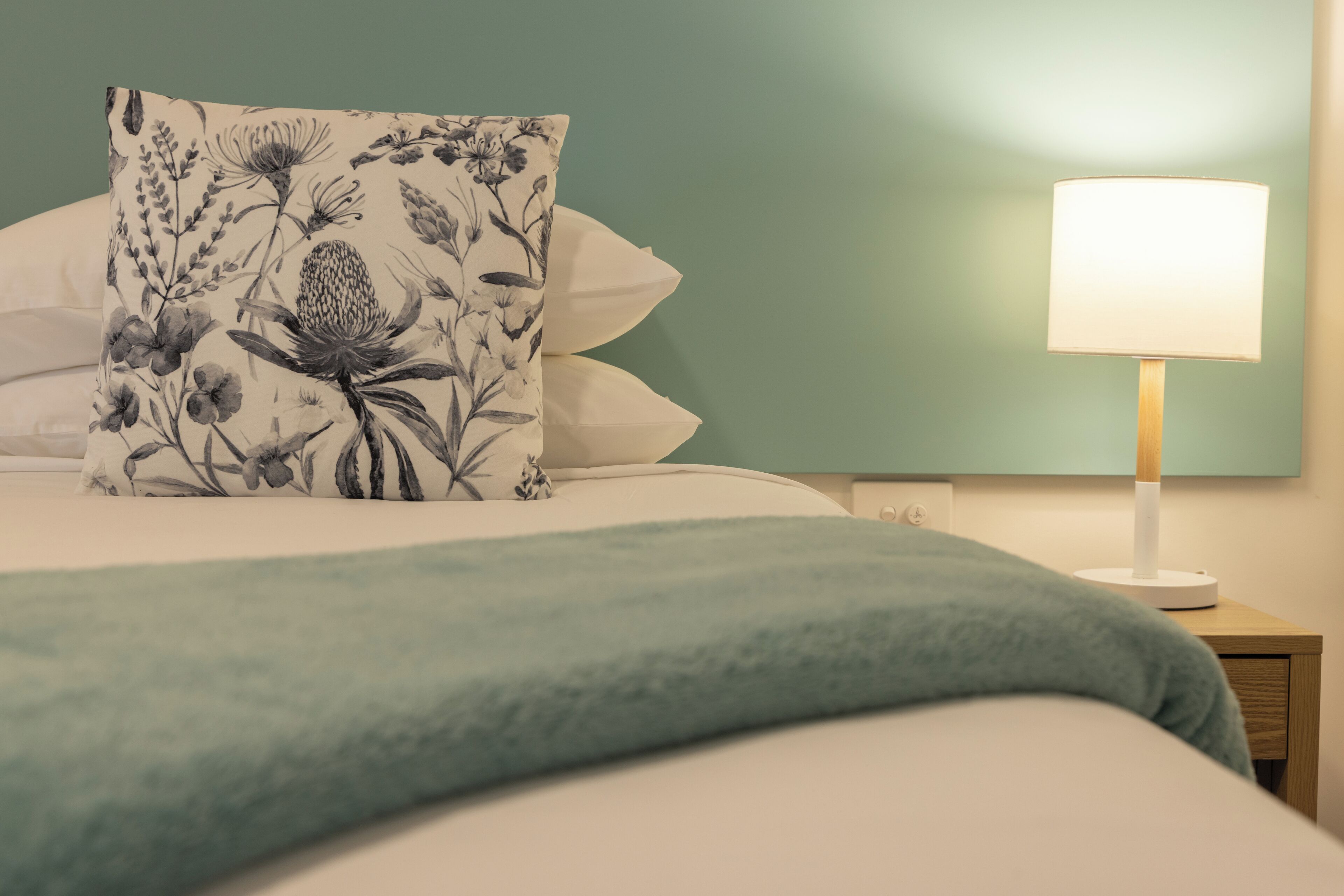 A neatly made bed with a floral pillow, a teal throw blanket, and a bedside lamp on a wooden nightstand.