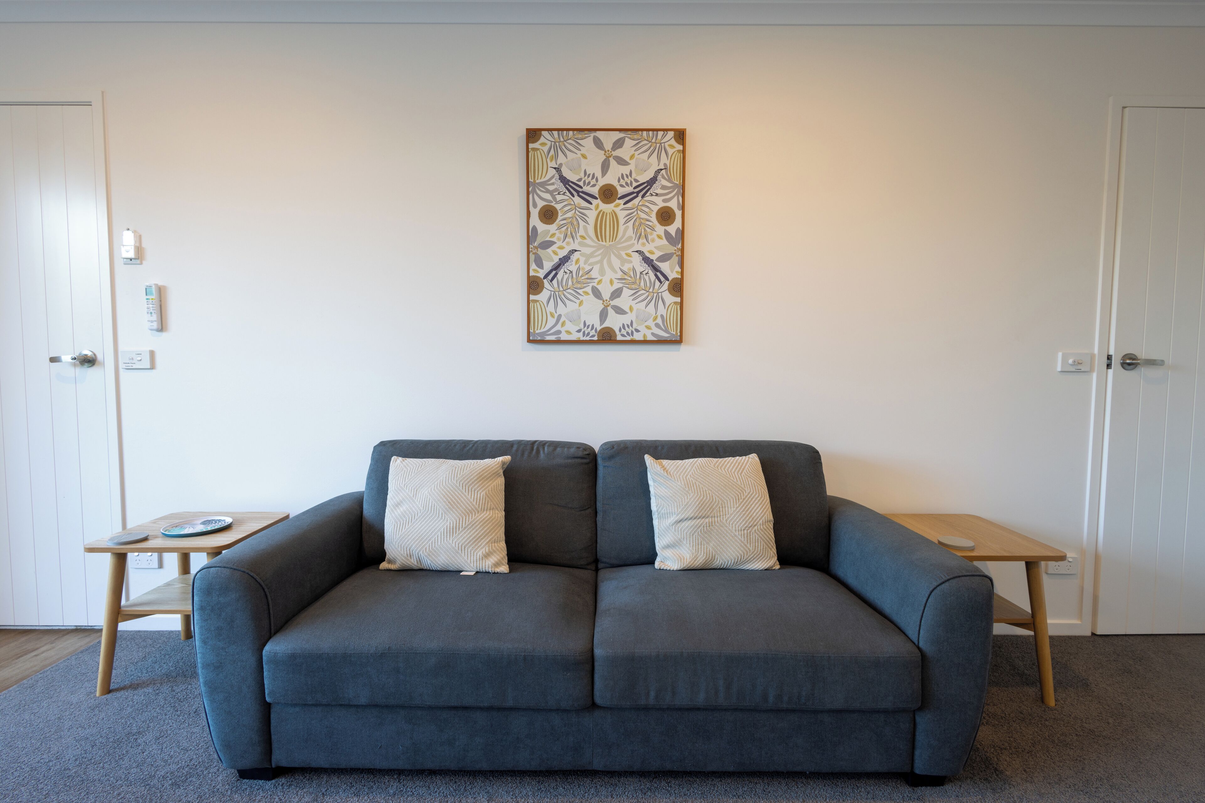 A modern living room with a grey sofa, two matching cushions, a wooden side table, a framed abstract artwork on the wall, and a white door in the background.