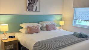 Un dormitorio con cama, dos mesitas de noche, un reloj de pared, un ventilador de techo, una ventana con persianas y un cuadro enmarcado en la pared.
