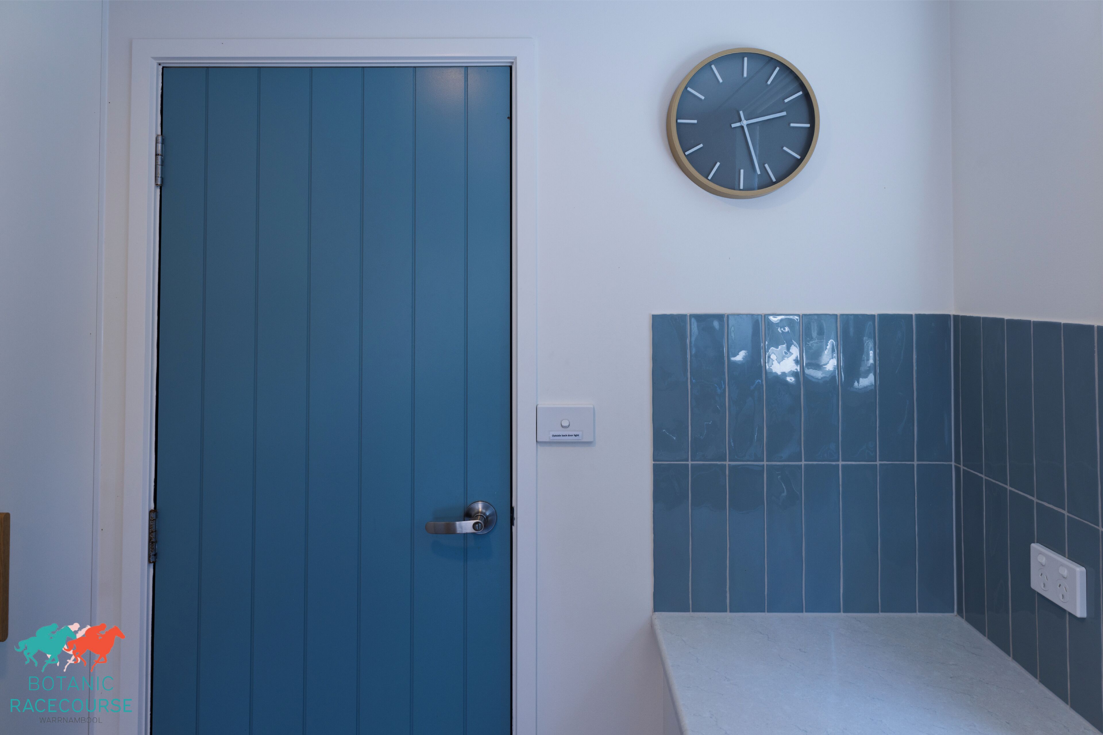 Un baño con puerta azul, un reloj en la pared y un salpicadero de azulejos.