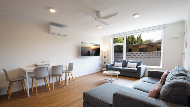 Una sala de estar moderna con un sofá, una mesa de comedor con sillas, un televisor montado en la pared y un ventilador de techo.
