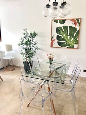 A modern dining area with a glass table and chairs, a potted plant, and a framed tropical leaf artwork on the wall.