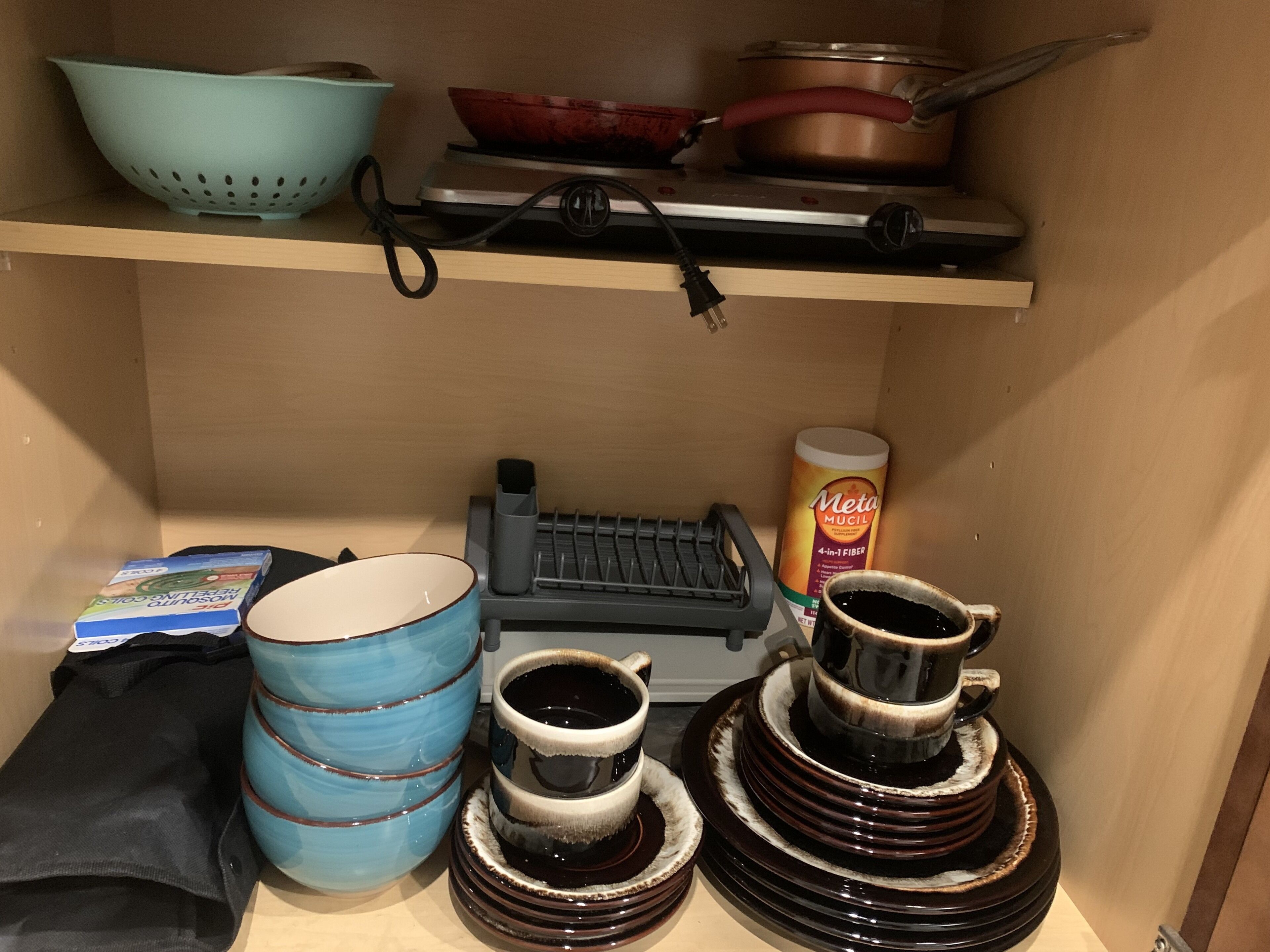 A kitchen shelf with a colander, a pot, a kettle, a coffee maker, and a can of Meta Mucil.