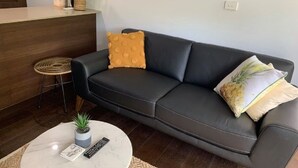 Ein modernes Wohnzimmer mit einem dunkelgrauen Ledersofa, einem runden Couchtisch mit Fernbedienung, einer kleinen Topfpflanze und einem gewebten Hocker.