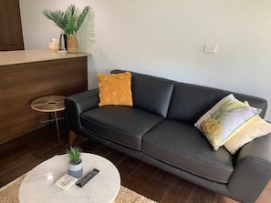 Un salon moderne avec un canapé en cuir foncé, une table basse ronde avec une télécommande, une petite plante en pot et un tabouret en osier.