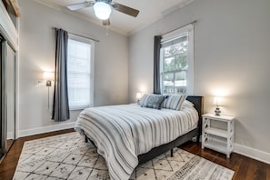 Une chambre à coucher avec un lit, une table de chevet, un ventilateur de plafond et une fenêtre avec des rideaux.