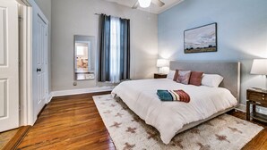Une chambre avec un grand lit, un ventilateur de plafond, une table de chevet, une lampe, une fenêtre avec des rideaux et un tableau au mur.