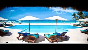A pool area with lounge chairs and umbrellas, a clear view of the ocean, and a sandy beach. - Cana Rock Chic Golf de Lux (Punta Cana)
