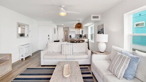 A modern living room with a white sofa, a wooden coffee table, and a kitchen in the background. - Beachfront Duplex | Shared Heated Pool | Pet Friendly (Gulf Shores)