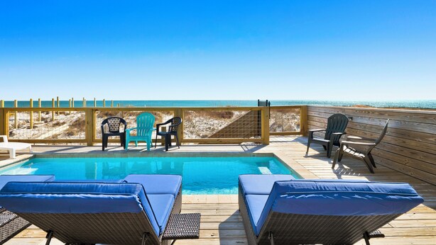 A wooden deck with a pool, lounge chairs, and a view of the beach.