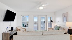 A living room with a flat-screen TV, a ceiling fan, a sofa, a coffee table, and a window with blinds.