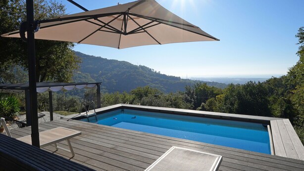 A pool with a wooden deck, a pergola with a canopy, and a view of a hilly landscape.