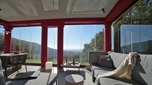 A modern outdoor seating area with a sofa and chairs, a round table, and a view of the landscape through large windows.
