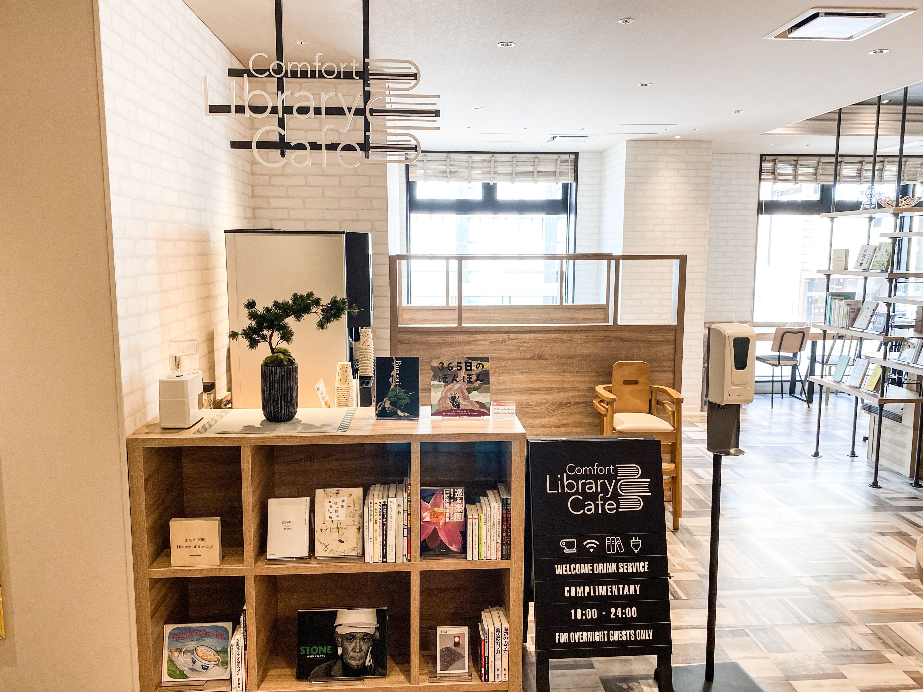 Ein Café in einer Bibliothek mit Bücherregalen, einer Kaffeemaschine und einem Schild, das einen kostenlosen Getränkeservice von 10:00 bis 24:00 für Übernachtungsgäste anzeigt.
