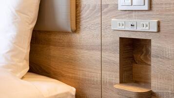 A wall-mounted control panel with a power outlet and USB port, a recessed shelf, and a bed with white bedding.