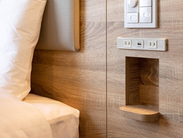 A wall-mounted control panel with a power outlet and USB port, a recessed shelf, and a bed with white bedding.
