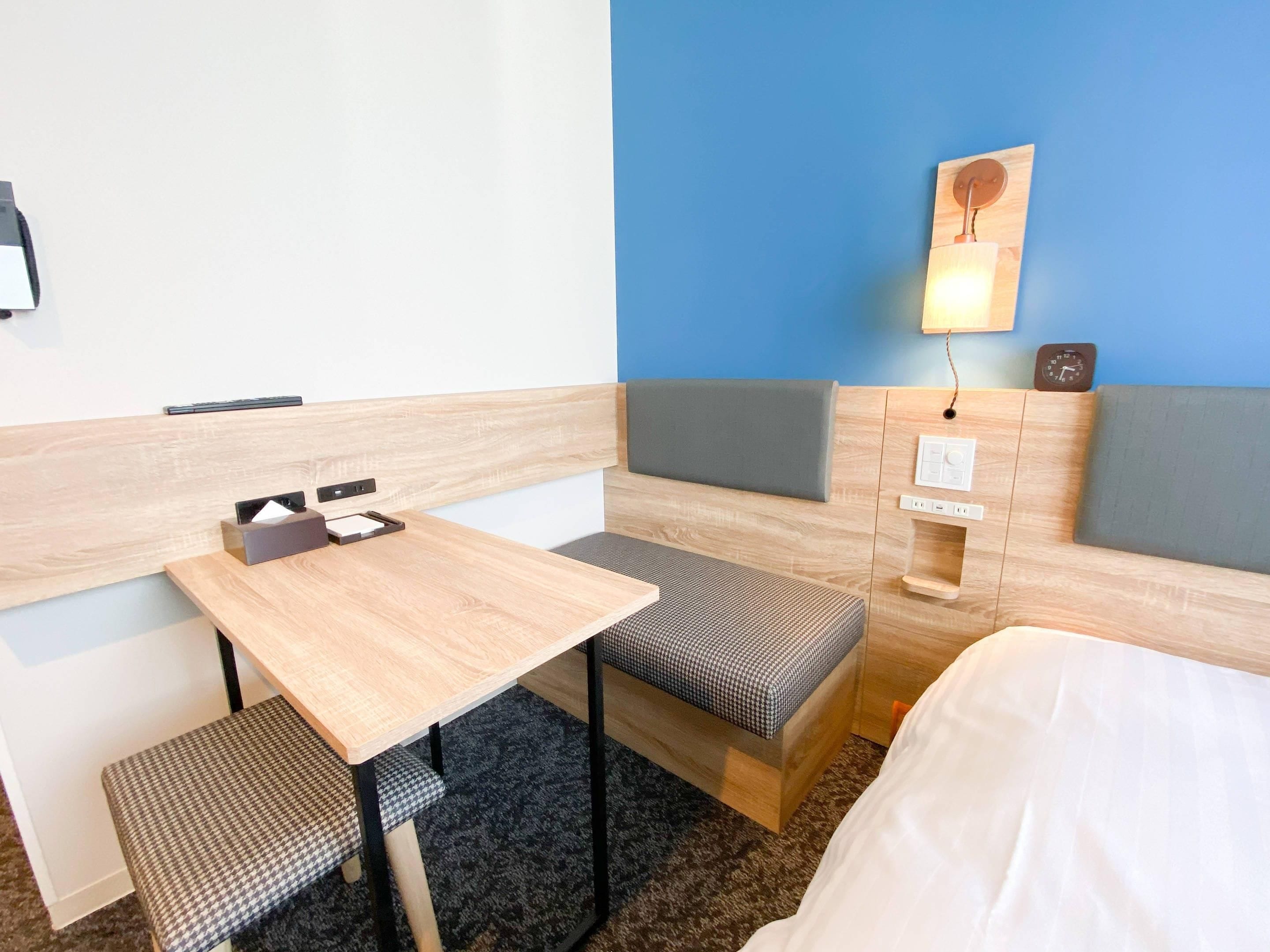 Hotellihuone, jossa on puinen työpöytä, kaksi pehmustettua istuinaluetta, sänky, seinään kiinnitetty valaisin ja kello.