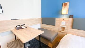 Hotellihuone, jossa on puinen työpöytä, kaksi pehmustettua istuinaluetta, sänky, seinään kiinnitetty valaisin ja kello.