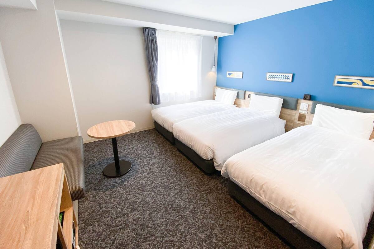 Habitación de hotel con dos camas, una mesita redonda pequeña, un sofá y una ventana con cortinas.